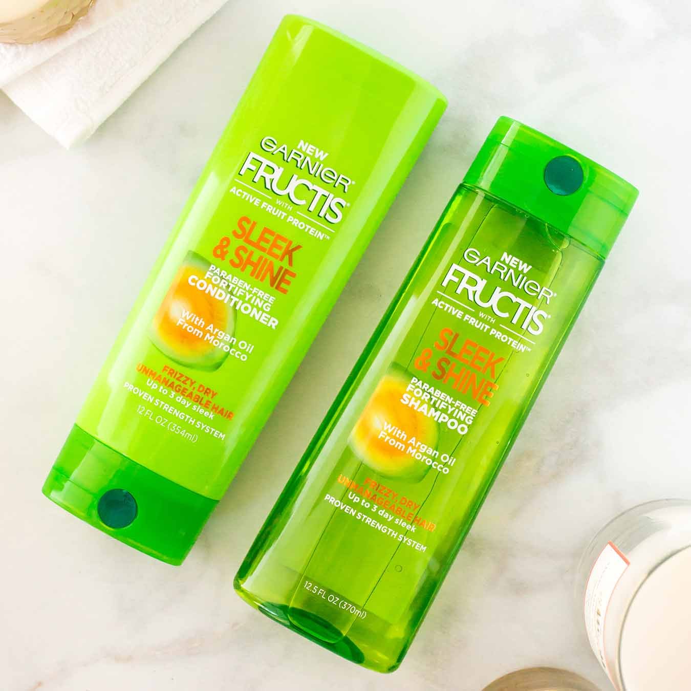 Garnier Fructis Sleek & Shine Shampoo and Fructis Sleek & Shine Conditioner on white marble next to three unlit white candles a white hand towel.